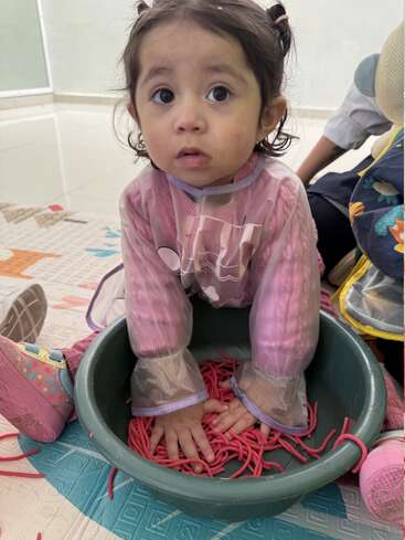 A young child with pigtails, wearing a pink smock, sits in a green tub filled with red noodles, exploring sensory play with wide-eyed curiosity.