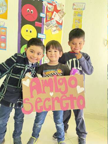 Three smiling boys pose together in a classroom, holding a cardboard sign that reads "Amigo Secreto" with hearts. Colorful educational posters decorate the cheerful background.