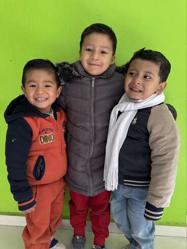 Three young boys stand smiling against a bright green wall. They're dressed warmly in jackets, scarves, and cozy clothes, radiating happiness and close friendship together.