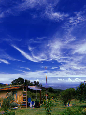 Uma pequena casa rústica com uma escada de madeira fica entre árvores verdes sob um céu azul vibrante cheio de nuvens finas, com vista para uma paisagem distante. Cena tranquila.