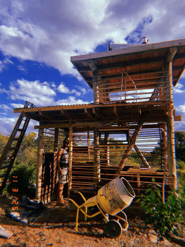 Uma pessoa está ao lado de uma estrutura de madeira parcialmente construída sob um céu azul com nuvens. Você pode ver um misturador de cimento amarelo e uma escada em primeiro plano.