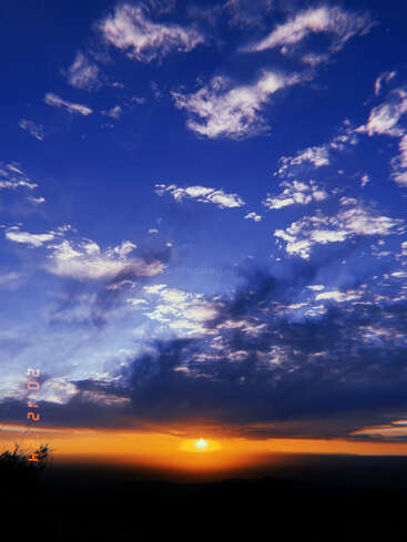 Um pôr do sol de tirar o fôlego, com tons vívidos de laranja e amarelo, encontra um céu azul profundo, pontilhado por nuvens dramáticas, criando uma atmosfera pacífica e encantadora.