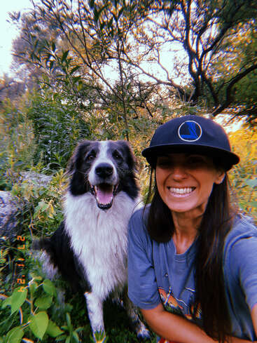 Uma mulher alegre com um boné preto sorri amplamente, posando ao ar livre com seu alegre cachorro preto e branco, cercada por uma vegetação exuberante e a luz do sol passando pelas árvores.