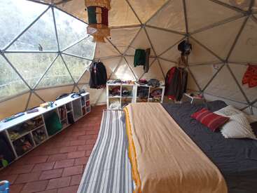 Um quarto aconchegante em forma de cúpula geodésica com uma cama grande, tapete listrado, prateleiras com cubículos, roupas penduradas e luz do sol que entra pelas janelas triangulares, criando uma atmosfera acolhedora.