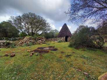 Eine traditionelle Steinhütte mit einem kegelförmigen Strohdach steht in einer grasbewachsenen, bewaldeten Gegend, umgeben von Felsen, Bäumen und einem bewölkten Himmel. Friedlich, urig.