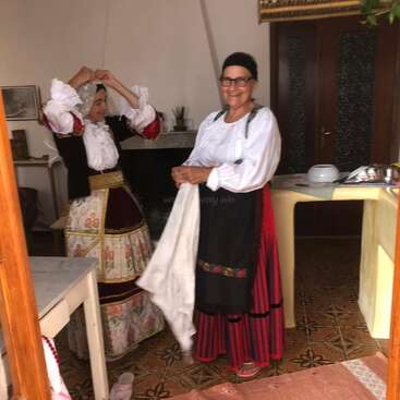 Zwei Frauen in traditionellen europäischen Trachten befinden sich in einem Raum. Die eine richtet ihr Kopftuch, während die andere lächelt und ein Tuch in der Hand hält. Der Raum ist gemütlich eingerichtet.