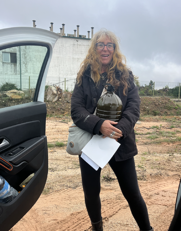 Uma mulher sorridente, com cabelos longos e ondulados e óculos, está ao ar livre, segurando uma garrafa grande e escura, papéis e uma bolsa cinza perto da porta aberta de um carro.