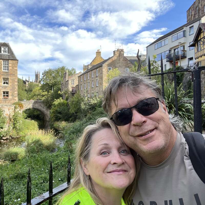 A smiling couple takes a close-up selfie outdoors, framed by historic stone buildings, greenery, a stone bridge, and a blue sky with scattered clouds.