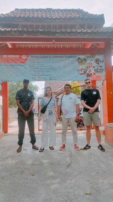 Vier Personen stehen vor einem Schultor mit einem großen Schild in indonesischer Sprache, unter einem Ziegeldach, und posieren für ein Gruppenfoto im Freien.