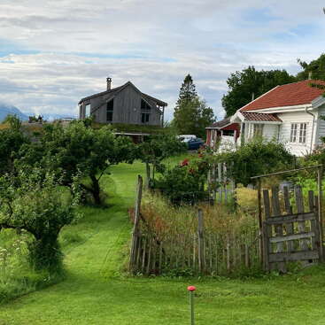 Uma cena rural tranquila mostra duas casas, um gramado verdejante, um jardim com cerca de madeira, macieiras, montanhas ao longe e um céu azul nublado acima.