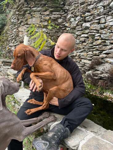 The image depicts a man sitting on a stone wall, cradling a brown dog in his lap, with another dog standing nearby, all set against a rustic stone wall backdrop.