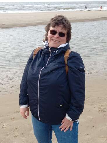 A woman wearing sunglasses, a navy jacket, and blue jeans stands smiling on a sandy beach with a body of water and ocean in the background.