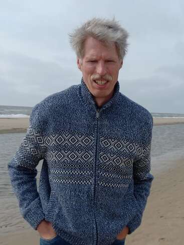 A man with gray hair and a mustache wearing a blue patterned sweater stands on a sandy beach with his hands in his pockets, smiling slightly.