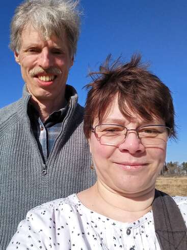 A middle-aged man and woman smile together outdoors on a sunny day, blue sky above, both dressed warmly, enjoying the bright weather and each other's company.