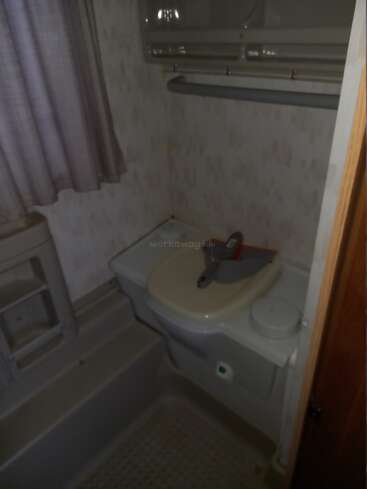 This image shows a small, dimly lit bathroom with a compact sink, a cleaning brush, plastic shelves, patterned wallpaper, purple curtains, and an overhead storage cabinet.