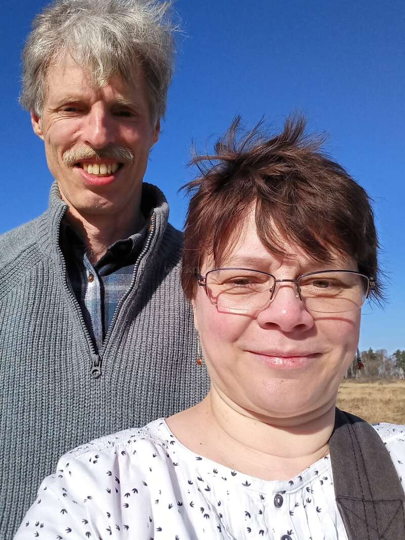 A middle-aged man and woman smile together outdoors on a sunny day, blue sky above, both dressed warmly, enjoying the bright weather and each other's company.