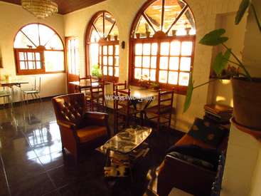 L'image représente une pièce aux murs blancs, aux fenêtres cintrées et au sol carrelé foncé, avec une table et des chaises, un canapé et une plante au premier plan.