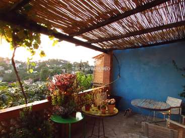 La imagen muestra un sereno balcón con techo de madera, con una pequeña mesa y sillas, rodeado de frondosas plantas y una pintoresca vista de una ciudad al fondo.