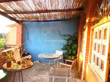 L'image représente un patio extérieur confortable, avec une petite table et des chaises, entouré de plantes et d'un mur bleu, avec un toit en bois et une porte sur la droite.