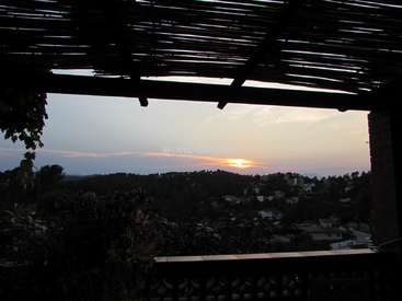 L'image représente un coucher de soleil serein depuis un balcon, avec une pergola en bois et une végétation luxuriante au premier plan, sur fond de ville à flanc de colline.