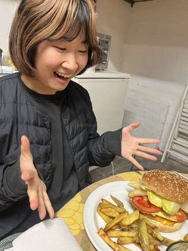 Ein junger Mensch guckt total begeistert auf einen Teller mit Essen, auf dem ein großer Burger mit Salat, Tomate, Käse und Pommes als Beilage liegt.