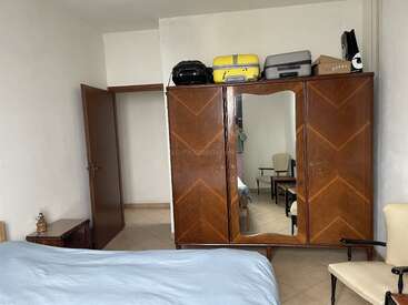 In diesem Zimmer gibt's einen Kleiderschrank aus Holz mit Spiegel, Koffer drauf, eine offene Tür, ein Bett mit blauer Bettdecke, Stühle und Fliesenboden.