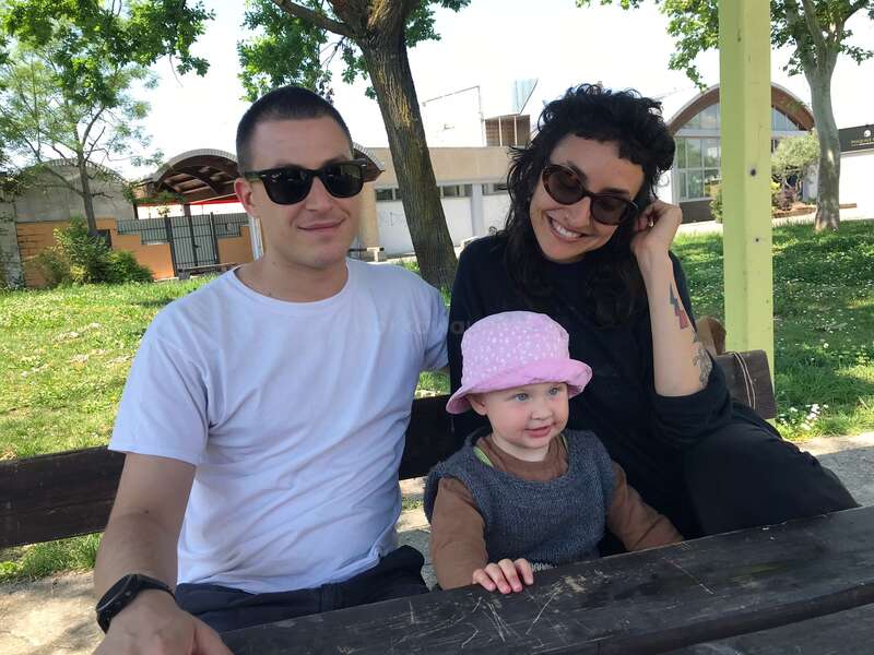 Eine fröhliche Familie sitzt auf einer Bank im Freien. Der Mann und die Frau tragen Sonnenbrillen, während das lächelnde Kind mit einem rosa Hut zwischen ihnen sitzt.