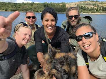 The image shows a group of five people posing for a selfie with a dog in front of a lake, surrounded by trees and hills under a blue sky.