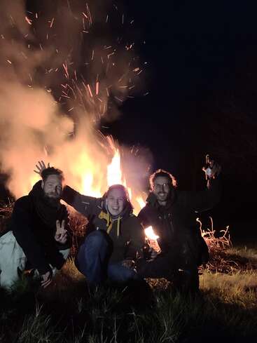 Three people sit in front of a large bonfire at night, with one holding a lit torch and another making a peace sign.