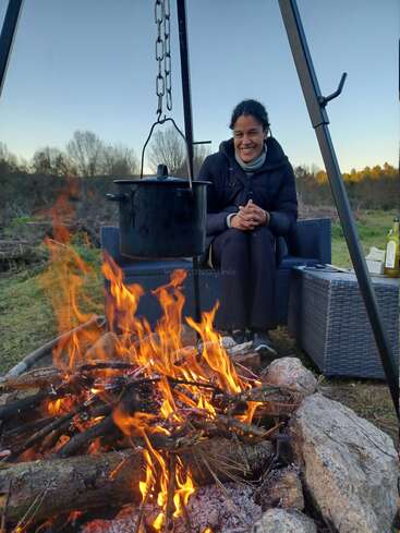 A woman sits on a couch beside a campfire, with a large pot suspended over the flames by a metal hook, in a serene outdoor setting.