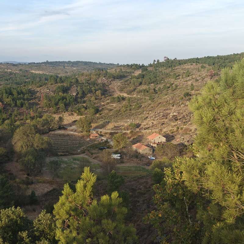 A scenic rural landscape with rolling hills, scattered trees, and a few rustic houses nestled in a valley. The view is peaceful, wide, and green.