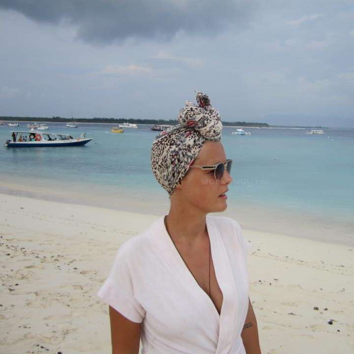 L'image représente une femme debout sur une plage, vêtue d'un haut blanc et d'un foulard, avec une vue sereine de l'océan et des bateaux en arrière-plan sous un ciel nuageux.