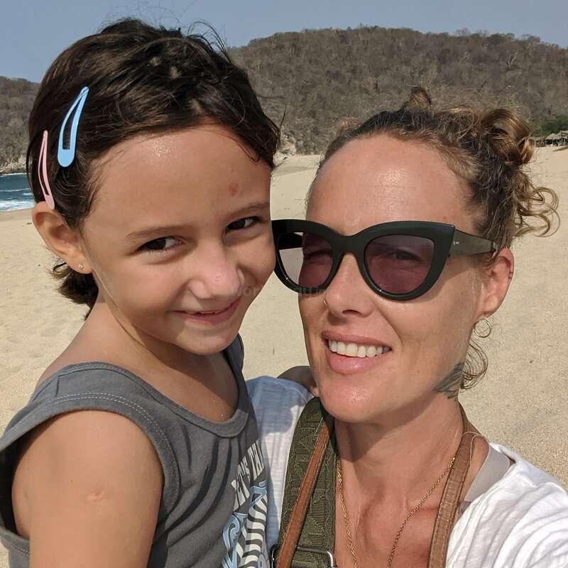 L'image montre une femme et une jeune fille posant pour un selfie sur une plage de sable, avec un plan d'eau et une zone boisée en arrière-plan.