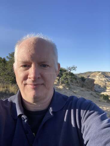 A man with short white hair is taking a selfie outdoors. He wears a blue shirt, with rocky hills, sparse trees, and clear blue sky behind him.