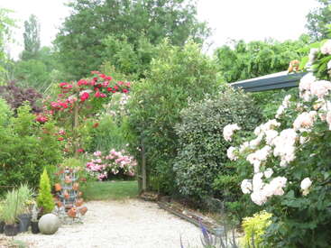 Um jardim exuberante repleto de vários arbustos verdes, rosas desabrochando e flores coloridas. Uma treliça coberta de rosas vermelhas fica perto de um caminho de cascalho.