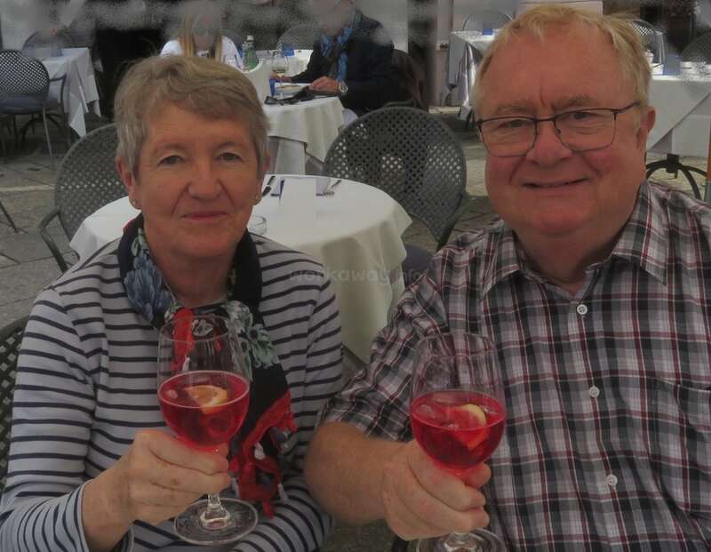 Um casal de idosos senta-se ao ar livre em um restaurante, sorrindo e segurando copos de bebidas vermelhas decoradas com frutas, desfrutando de um momento agradável juntos.