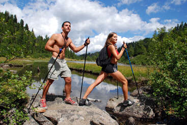 Ein fitter Mann und eine Frau wandern mit Wanderstöcken an einem malerischen Fluss entlang, umgeben von einem üppig grünen Wald und blauem Himmel mit vereinzelten Wolken. Outdoor-Abenteuer.