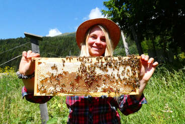Eine lächelnde Frau mit Strohhut und kariertem Hemd steht draußen in einer sonnigen, grasbewachsenen Berglandschaft und hält einen Bienenstock voller Bienen in der Hand.