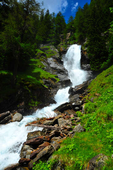 Ein mächtiger Wasserfall rauscht über felsiges Gelände, umgeben von üppig grünen Bäumen und lebhafter Vegetation unter einem strahlend blauen Himmel mit vereinzelten Wolken. Eine ruhige Naturlandschaft.