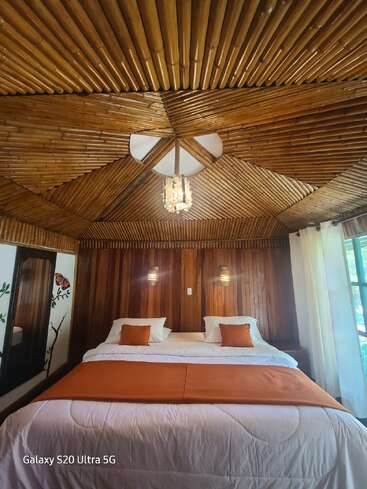 Este quarto aconchegante apresenta um teto de bambu, paredes de madeira, uma cama grande com detalhes em laranja, iluminação suave, um espelho e luz solar natural através das cortinas.