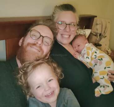 Uma família feliz de quatro pessoas posa para uma selfie em uma cama, os pais sorrindo com seus dois filhos pequenos, incluindo um bebê de pijama estampado.