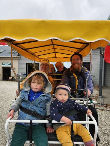 A family of six, including two young children, rides together on a covered four-wheel bike with a bright yellow canopy, smiling and enjoying their outing.