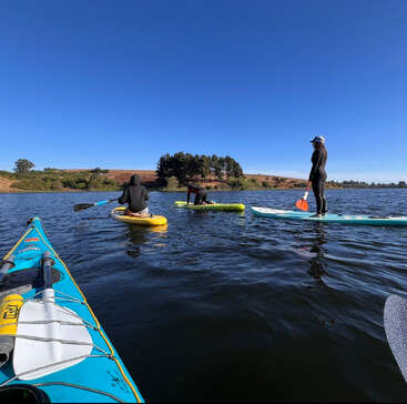 Four people enjoy paddleboarding and kayaking on a calm lake under a clear blue sky, surrounded by green trees and distant hills on a sunny day.