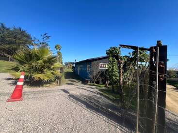 A gravel driveway leads to a blue house surrounded by greenery and trees. A red traffic cone and wire gate are visible under a clear blue sky.
