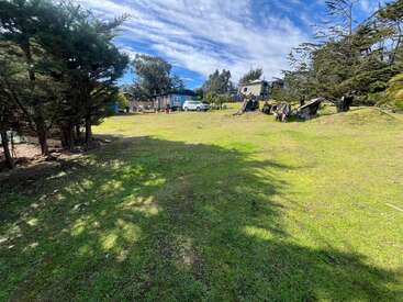 A grassy open field surrounded by trees, with a few houses and vehicles in the background under a blue sky with scattered clouds, bathed in sunlight.