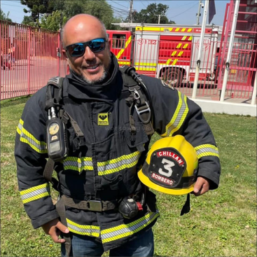 A smiling firefighter wearing a black uniform with yellow stripes, holds a yellow helmet labeled "Chillan Bombero 3", standing beside a firetruck and red gate outdoors.
