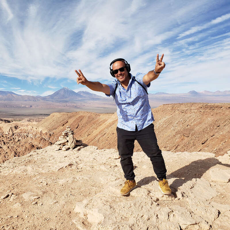 A man wearing headphones and sunglasses stands on a rocky desert landscape, flashing peace signs with both hands, smiling under a bright, expansive sky.