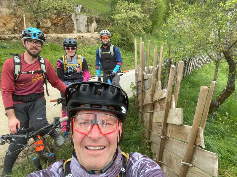 The image shows four mountain bikers posing for a selfie on a trail, with one person in the foreground and three behind, all wearing helmets and colorful clothing.