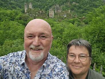 A smiling couple takes a selfie in front of lush green scenery with impressive ancient castle ruins visible on a hillside in the background. Beautiful moment.