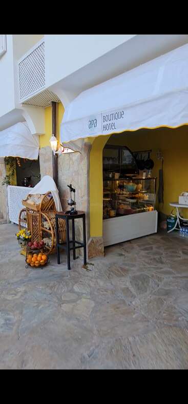 Esta imagen muestra la entrada del Apa Boutique Hotel con un acogedor expositor exterior, cestas de fruta, una lámpara rústica y un mostrador de pastelería en el interior, de ambiente acogedor.